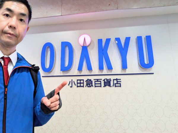 町田小田急百貨店のロゴ前で人差し指を立てる男性の手相占い師セルフィー。