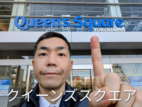 クイーンズスクエア横浜の入口前で人差し指を立てる手相占い師・後藤のセルフィー。オーバーレイテキストは「クイーンズスクエア」。